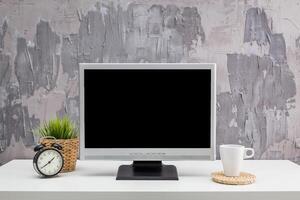 Desk setup featuring a blank computer monitor, clock, plant, and cup in a modern workspace with textured wall photo
