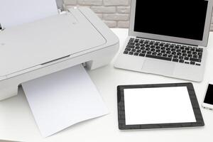 Printer in an office setting with a laptop, tablet, and smartphone on a desk during the day photo