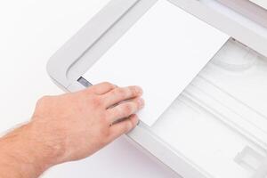 Hand placing a blank sheet of paper into a scanner on a white surface in a well-lit room during daytime photo