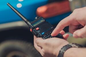 Individual operates handheld radio while standing next to blue vehicle in an outdoor setting during daytime photo