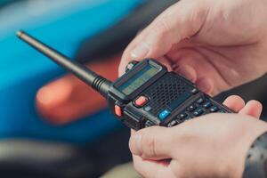 Hands hold a two-way radio while a blue vehicle is in the background during a sunny day photo