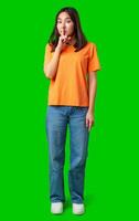 Young woman in orange shirt holding finger to lips on bright green background while signaling to keep quiet and maintain secrecy photo