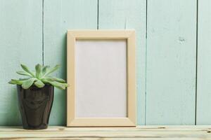 Decorative plant and empty picture frame on wooden table with teal background photo