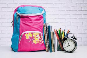 Brightly colored backpack and school supplies on a table with a clock in a well-lit study space photo