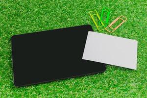 Workspace setup with tablet, blank card, and colorful paper clips on green grass background photo