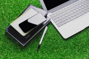 Work setup on artificial grass with laptop, smartphone, notepad, and pen during a sunny day photo