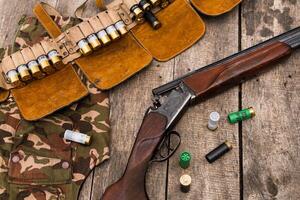 Hunting equipment displayed on a wooden table with shells and camouflage attire in a natural setting photo
