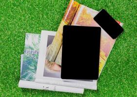 Digital and print media table arrangement with magazines, tablet, and smartphone on green grass backdrop photo