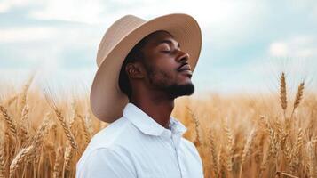 A man stands peacefully in a golden wheat field, wearing a wide-brimmed hat. He closes his eyes, basking in the warm sunlight and the gentle breeze of a tranquil afternoon photo