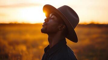 A man stands peacefully in a golden field as the sun sets behind him, wearing a stylish hat. He appears lost in thought, embracing the tranquility of the moment photo