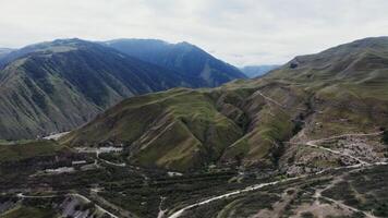 A view of the mountain valley, overgrown with grass and forest, many winding roads run along the rocky slopes. The beauty of the Caucasus mountainous nature from a bird's eye view from a drone photo