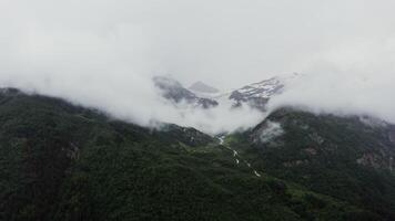 A bird's eye view of mountain valley with narrow, branched mountain river flowing between dense forest in lowland between the mountains, at the foot of rocky mountains covered with snow and clouds photo