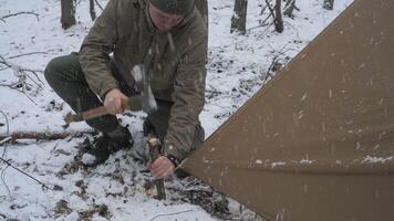 un persona es asegurando un tienda estaca dentro el Nevado suelo utilizando un hacha. el rodeando zona caracteristicas cubierto de nieve árboles, creando un sereno invierno cámping ambiente. video