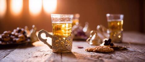 Wooden table set with a cup of tea in an ornate holder, a plate of dates, and a small bowl of dates. Soft, warm lighting and additional cups of tea. photo