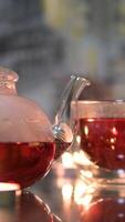 a woman's hand adjusts candles on a table cups of red tea and glass teapot sit on a stand, warmed by a candle underneath. One cup is lifted to take a sip. video