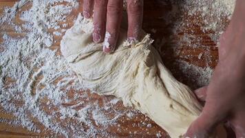 Close-up of hands kneading raw dough on a floured wooden surface, stretching and pulling elastic dough with visible texture, rustic home baking process in warm natural lighting video