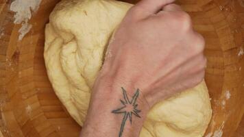 Close-up of tattooed male hands kneading dough in a wooden bowl, fingers covered in flour, veins visible, rustic wooden surface, warm and soft kitchen lighting creating a cozy and homely baking video