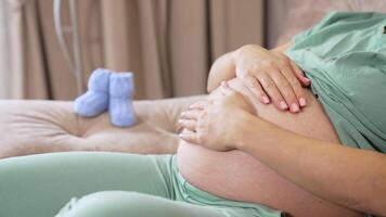 Caring future mother stroking her big tummy while resting on the sofa. Woman caressing her child in the big belly. Two little baby socks are on the armrest at he backdrop. video