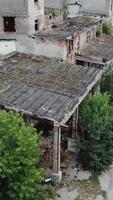 Aerial view. Ruined old building, tilt down to the dry grass and bushes. Abandoned factory halls with broken glass on windows. Apocalyptic scene with destroyed warehouse. Vertical video