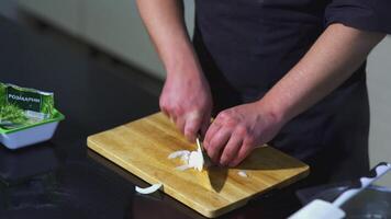 The cook cuts the onion. ART. Man in the kitchen cuts white onion into half rings with knife. Next to the board is spatula and bag of rosemary video