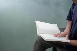 Boy opening blank book sitting on wooden bench at lakeside on water surface background,concept of education and work,blank notebook copy space for design,Hand opening blank book,boy holding notebook. photo