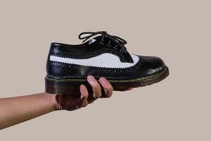 A man's hand holds a black and white brogue wingtip shoe with a rubber outsole made from genuine cowhide. Men's hands holding elegant and shiny two tone shoes on a cream background photo