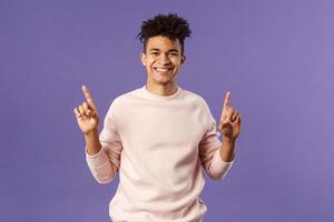 Waist-up portrait of joyful good-looking hispanic man with dreads, promoting product or company banner hanging on top, pointing fingers up, smiling satisfied, recommend subscribe or click link photo