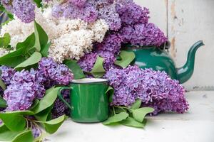A large bunch of lilacs in different shades are arranged around a green enamel mug and teapot on a white rustic table, creating a charming still life scene, photo