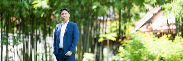 Asian handsome glasses man in a blue suit and the same color pants is looking at the camera, smart smiling and standing in the garden. photo