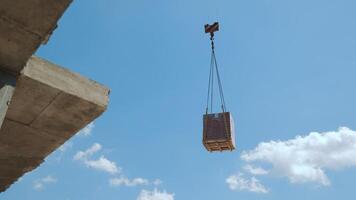Crane lifting pallet of bricks at construction site, A crane lifting a pallet of bricks high into the air at a construction site, set against a clear blue sky with some clouds video