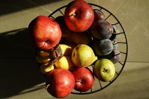 A fruit bowl that is full of bananas,apples, and kiwi on a countertop. photo