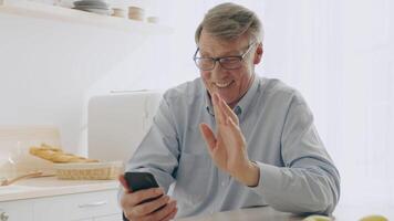 A senior man in his 60s enjoys a call in a sunny kitchen. He smiles and waves at the screen, exuding warmth and happiness in this relaxed home environment. photo