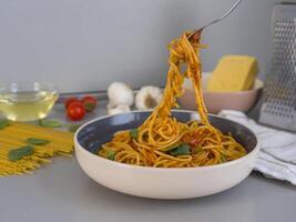 Balanesi pasta with tomato sauce and basil is being lifted by a fork from a bowl, with fresh ingredients like tomatoes, garlic, olive oil and cheese visible on a kitchen counter photo