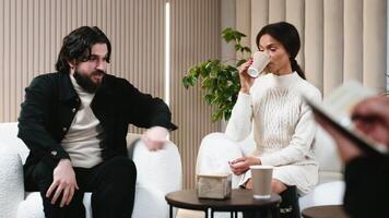 A couple sits on a grey couch in a therapy session, looking down with expressions of sadness or worry. A therapists hand is visible in the foreground, holding a pen and notepad video
