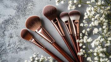 Five high-quality makeup brushes in varying sizes lay together, showcasing their soft bristles and polished wooden handles, surrounded by small white flowers against a textured gray surface photo