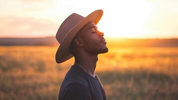 A man stands peacefully in a golden field during sunset. Wearing a wide-brimmed hat, he closes his eyes, embracing the calming atmosphere and warm light that surrounds him photo
