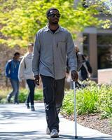A man with sunglasses confidently walks with a cane on a sunlit path, surrounded by trees and people, depicting independence. photo
