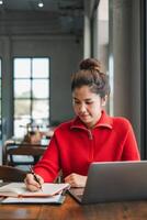A focused young woman in a red sweater writes in a notebook while using a laptop in a bright, modern cafe setting. photo