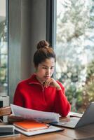 Woman in red sweater concentrating on work at home office, using laptop and reviewing documents by a large window. photo