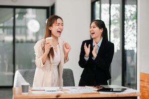 Two businesswomen in a modern office, laughing and celebrating success with documents and coffee on the table. photo