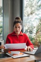 Woman in a red sweater working on a laptop and reviewing documents at a sunlit table by a window. photo