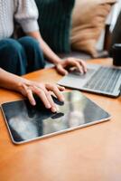 Hands working on a laptop and tablet on a wooden table, surrounded by cushions, in a cozy home environment. photo