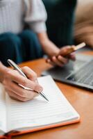 Close-up of a person taking notes in a notebook with a pen, holding a smartphone, and using a laptop on a wooden table. photo