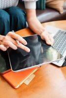 Close-up of hands using a digital tablet with a stylus, next to a laptop, on a wooden table in a casual environment. photo