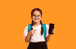Asian Schoolgirl Showing Smartphone Empty Screen Recommending Educational App Smiling To Camera Posing Over Yellow Studio Background photo