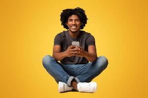 Great App. Cheerful young black guy sitting on floor with smartphone in hands, happy african american man browsing new application or messaging with friends over beige studio background, copy space photo