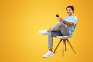 Portrait of excited smiling handsome millennial guy sitting on chair, using mobile application on modern smartphone, chatting with friends or watching funny , orange background, vertical photo