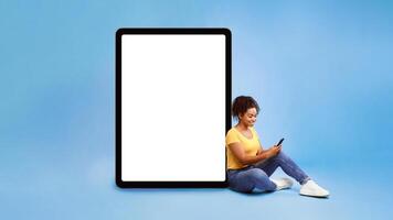 Full length of young African American lady using cellphone, sitting next to giant tablet pc with empty white screen, blue background. Mockup for app or website, space for advertisement design photo