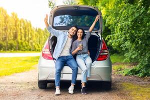 Carefree multiracial millennial couple having break at countryside while car trip, sitting on open trunk, drinking coffee, embracing and cheerfully smiling at camera, raising hands up, free space photo