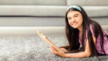 Domestic hobbies concept. Cheerful Indian teenage girl reading exciting book, lying on soft carpet at home. Lovely adolescent enjoying captivating story on weekend morning photo